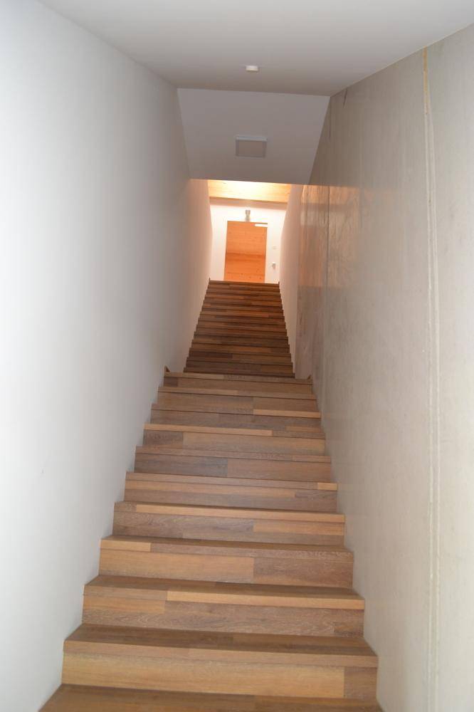 Eine lange Holztreppe mit hellen Stufen und weißen Wänden führt nach oben zu einem Flur.