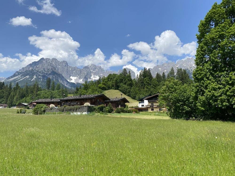 Weitläufige grüne Wiese mit traditionellen Häusern und beeindruckendem Bergpanorama unter strahlend blauem Himmel.