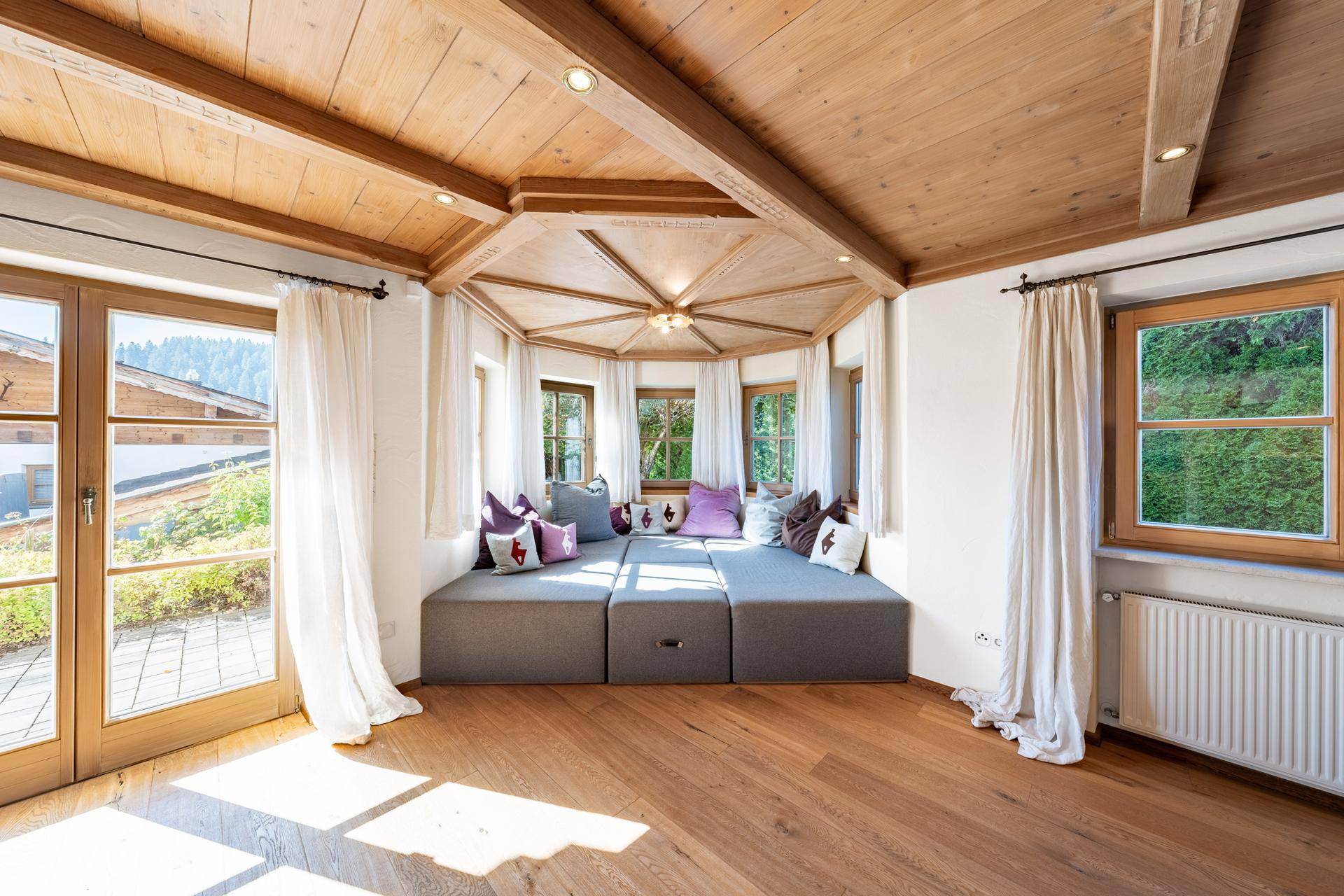 Gemütliche Fenstersitzbank mit Kissen in einem lichtdurchfluteten Raum mit Holzbalkendecke und großen Fenstern.