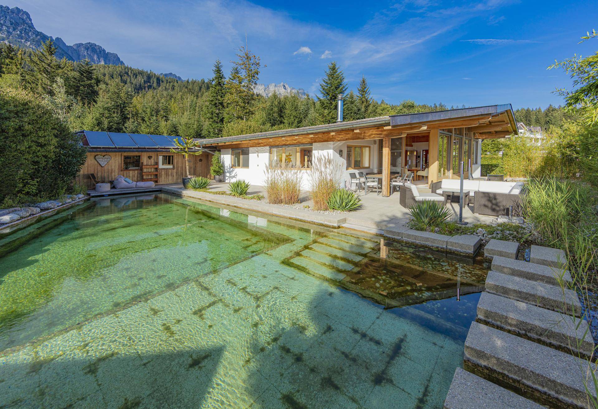 Modernes Haus mit Naturpool, großer Terrasse, Bergblick und Photovoltaikanlage in grüner Umgebung.