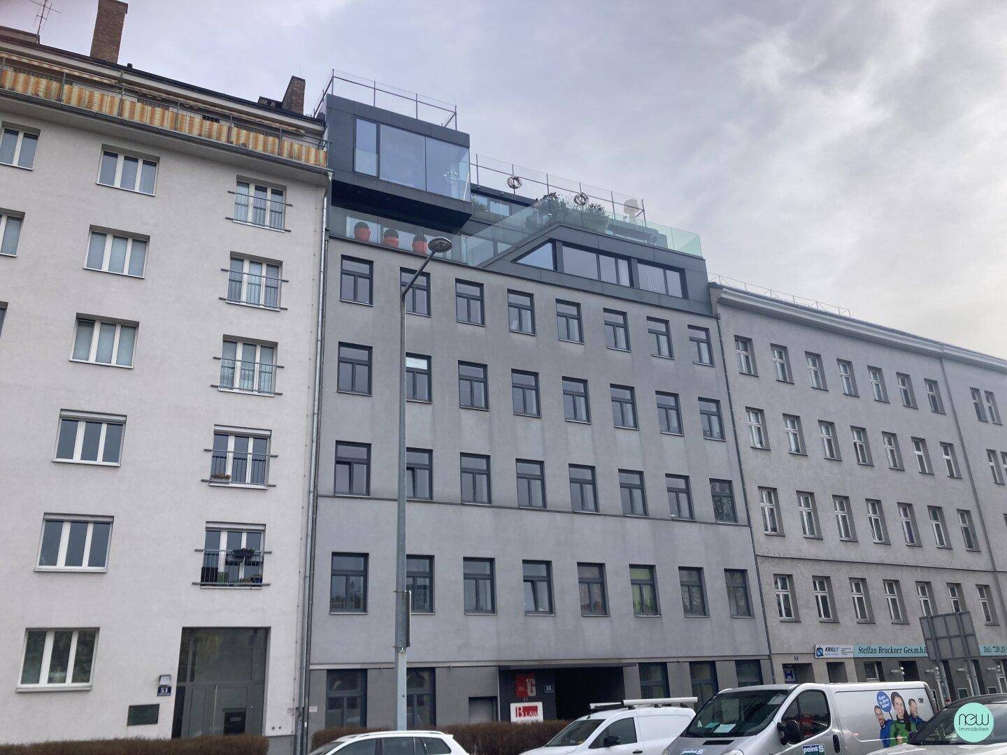 Moderne Gebäudefassade mit mehreren Etagen und einem gläsernen Dachaufbau in urbaner Umgebung.