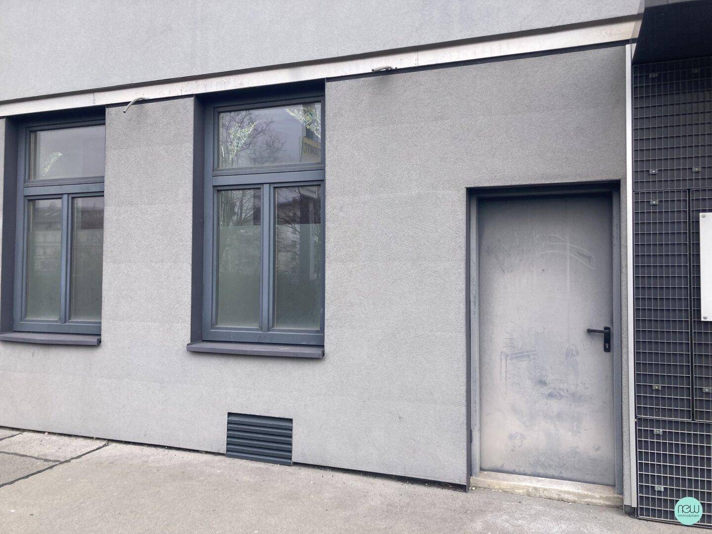 Graue Gebäudefassade im Erdgeschoss mit zwei Fenstern und einer einfachen Metalltür, die zur Straße führt.