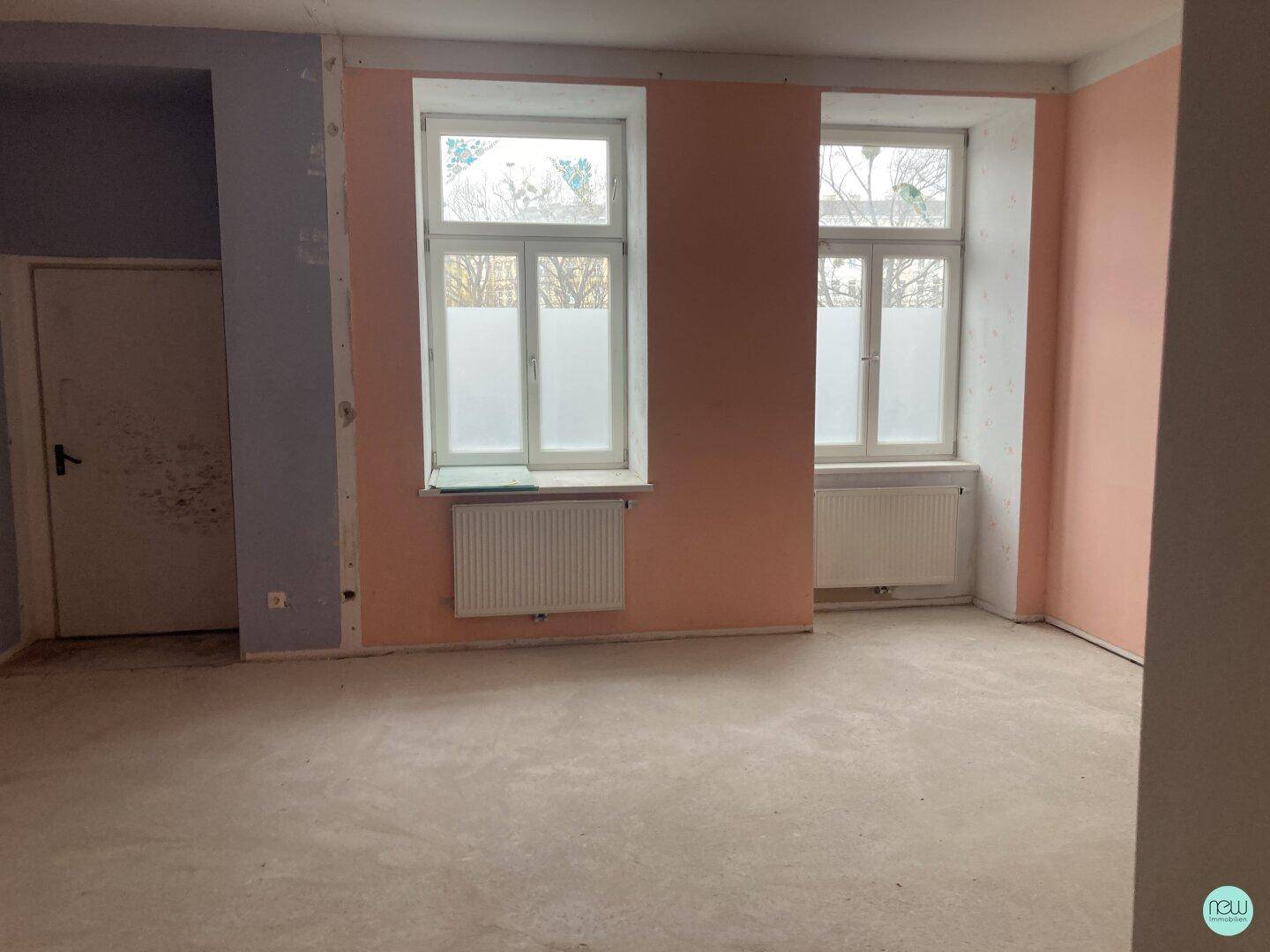 Renovierungsbedürftiger Raum mit zwei Fenstern, Heizkörpern und Wänden in verschiedenen Farbtönen.