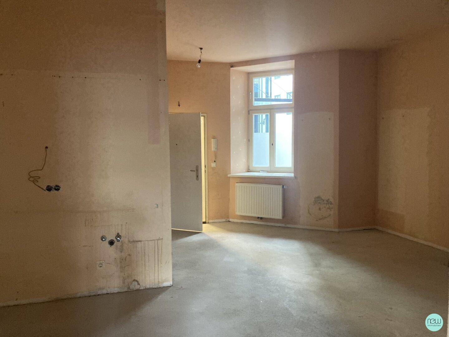 Unfertiger Innenraum mit einem Fenster, Heizkörper und sichtbaren Elektroinstallationen an der Wand.