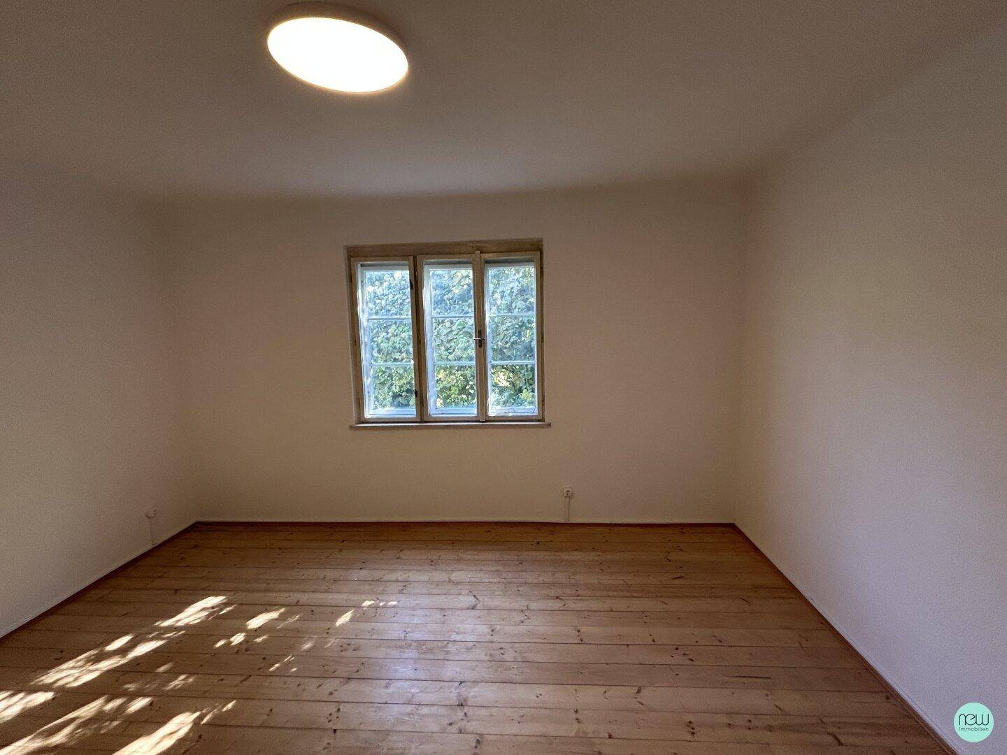 Helles, leeres Zimmer mit Holzdielenboden und einem Fenster mit Blick ins Grüne.