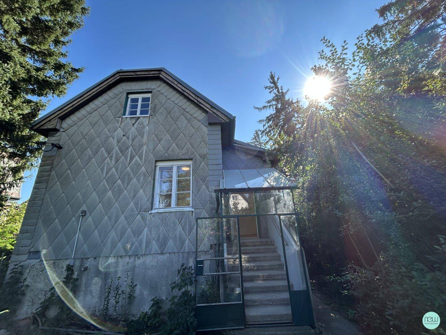 Seitenansicht eines Hauses mit Schieferfassade, Außentreppe und umgebender Natur bei Sonnenschein.