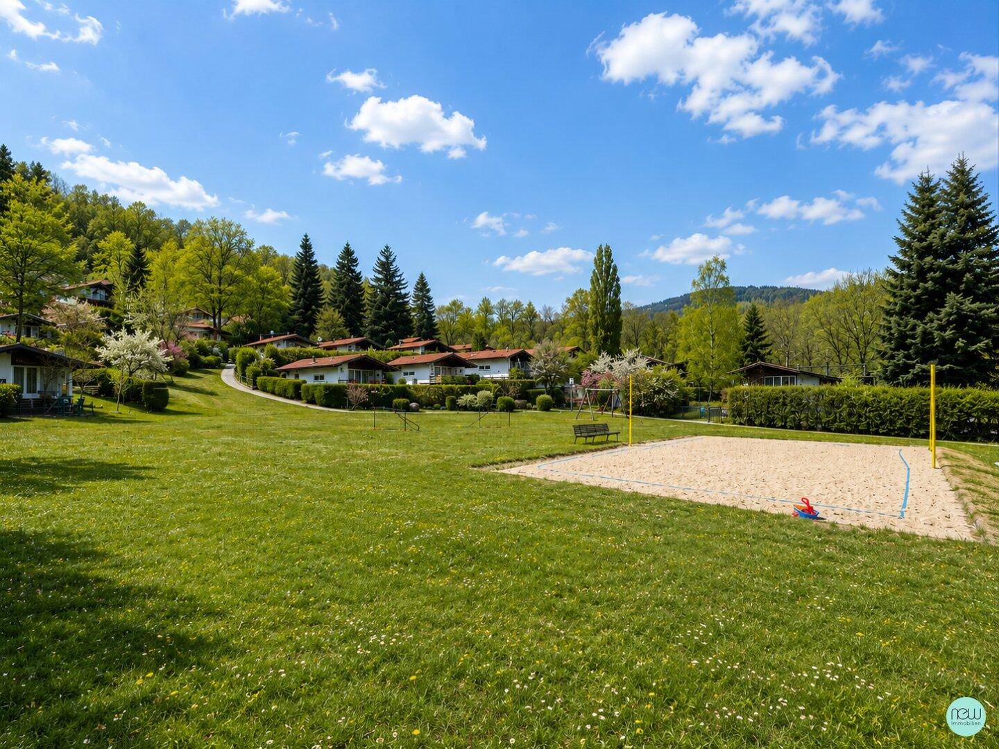 Große Rasenfläche mit Sandvolleyballfeld und Spielplatz, umgeben von Häusern und Natur.