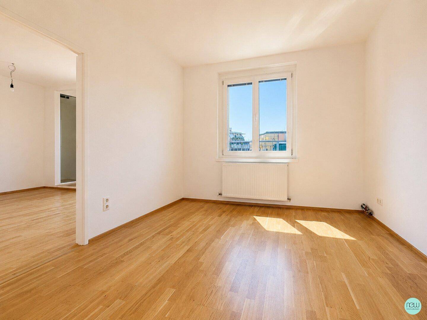 Geräumiges, helles Zimmer mit Parkettboden, weißen Wänden und einem großen Fenster für viel Tageslicht.