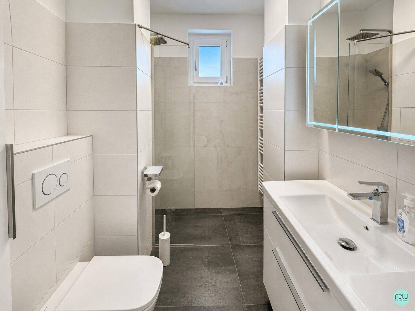Modernes Badezimmer mit hellen Fliesen, ebenerdiger Dusche, wandhängendem WC und Waschtisch mit Spiegelschrank.