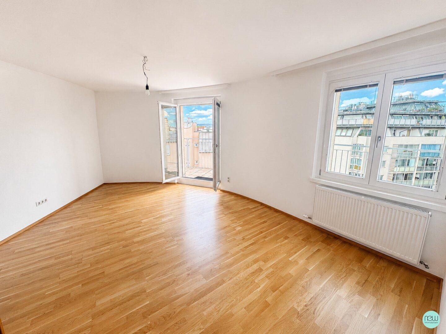 Helles, leeres Zimmer mit Parkettboden, weißen Wänden und Zugang zu einem Balkon.