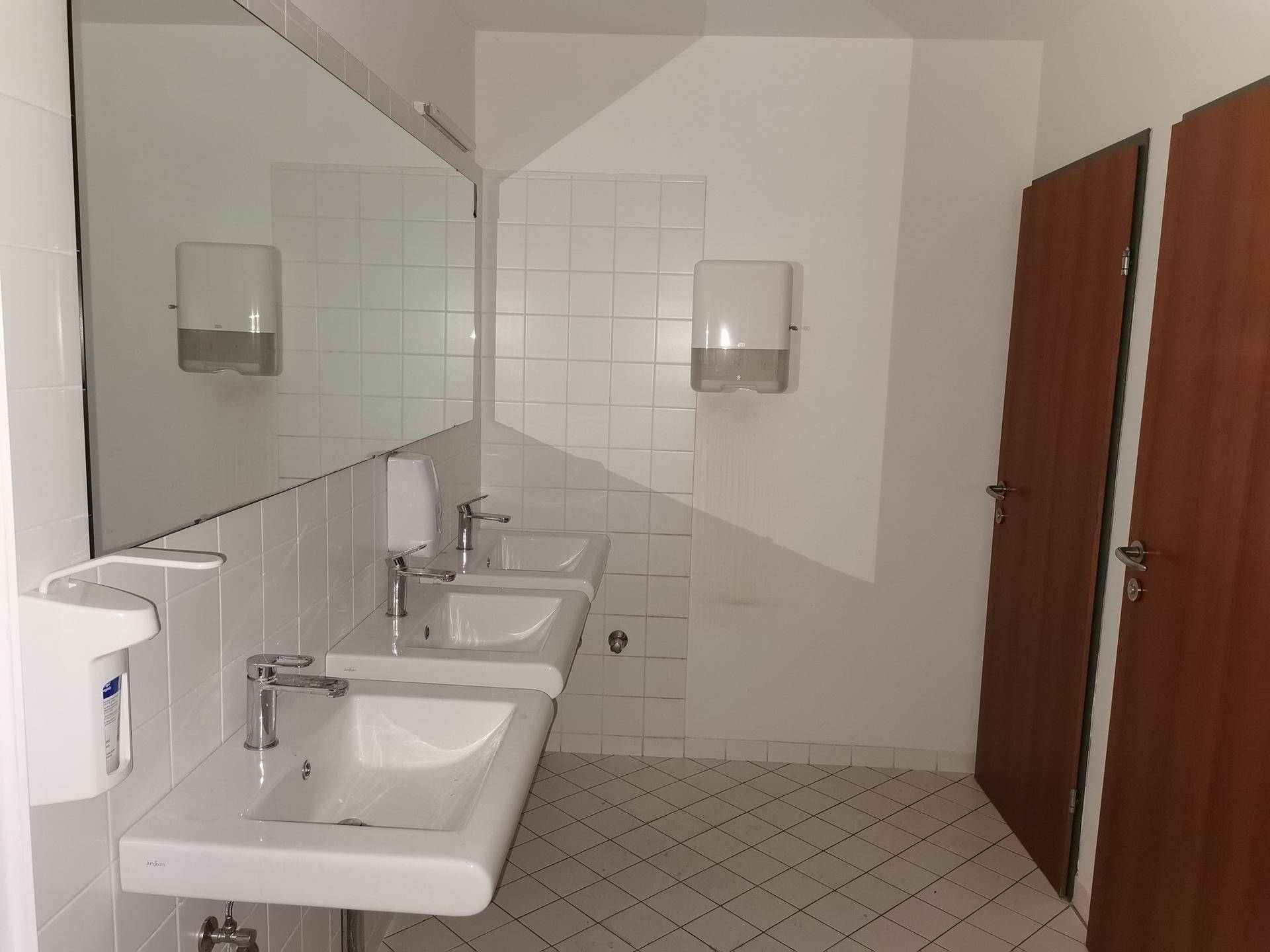 Funktionaler Sanitärbereich mit mehreren Waschbecken, großen Spiegeln und weißen Fliesen.