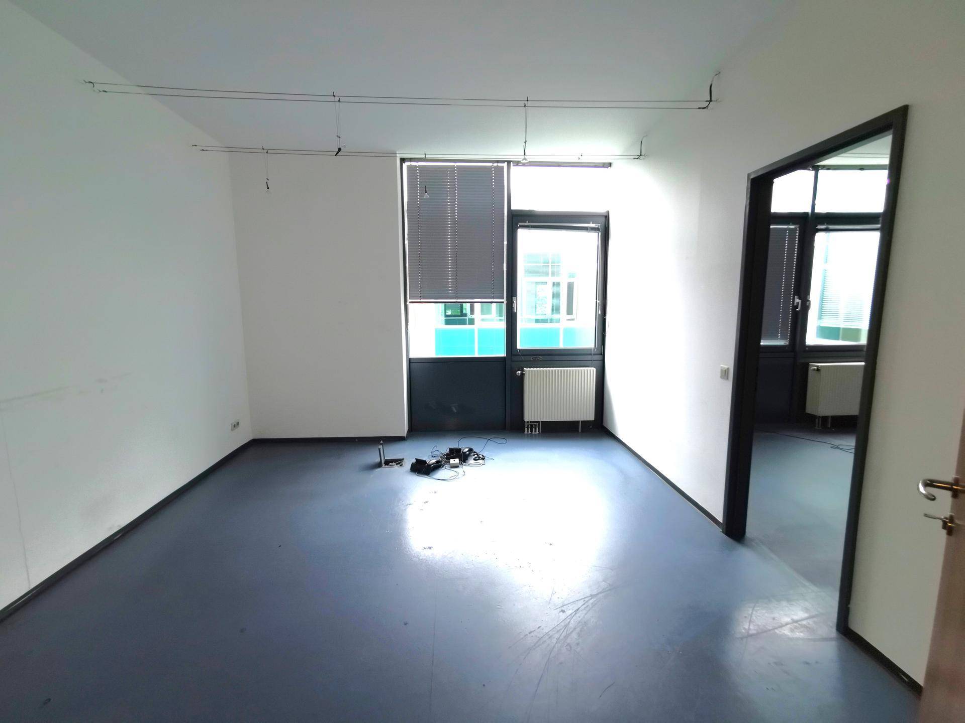 Leerer Büroraum mit blauem Vinylboden, Heizkörper und Fenster mit Jalousien.
