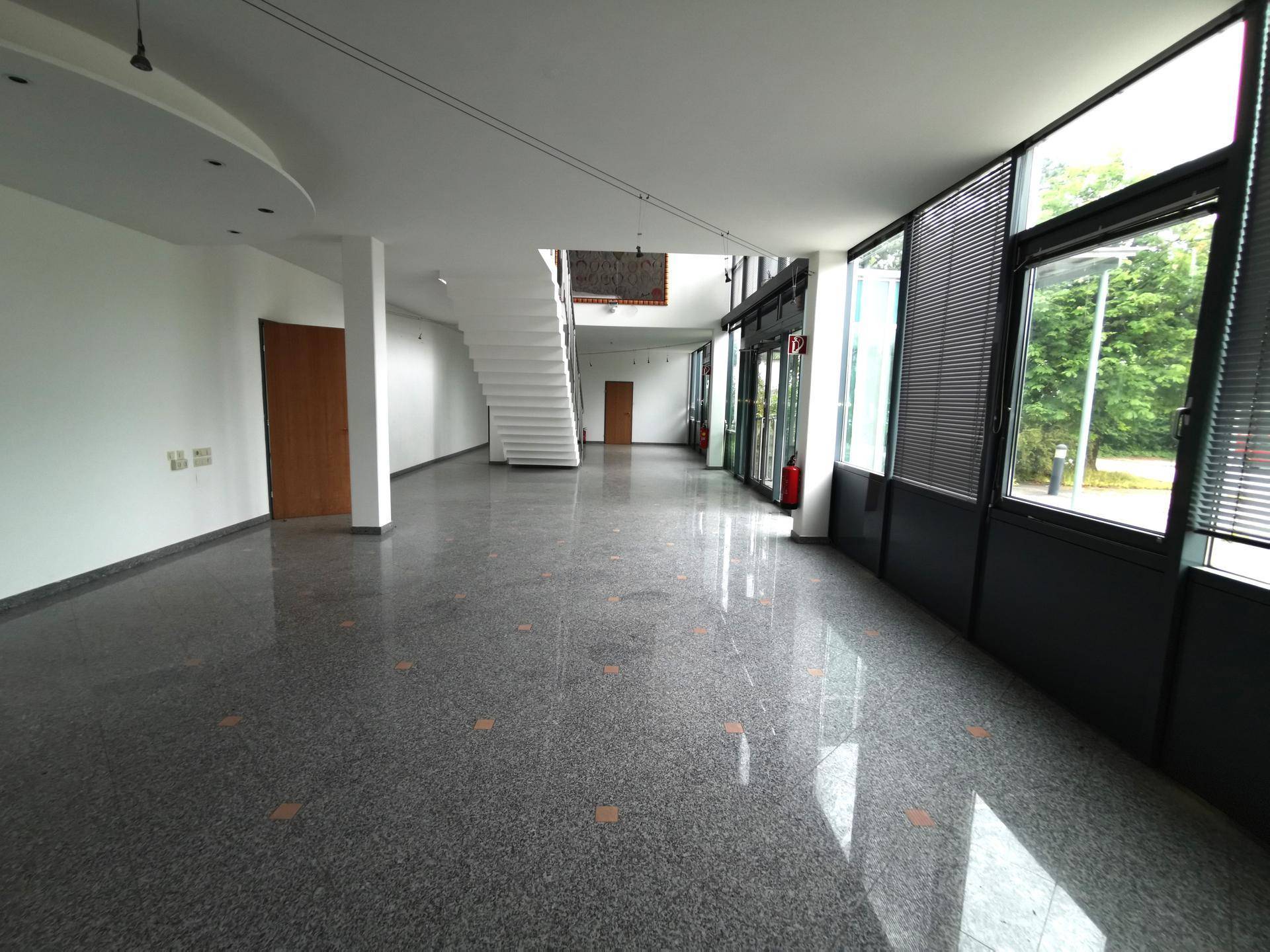 Weitläufiger Eingangsbereich mit glänzendem Terrazzoboden, Treppe und großen Fensterfronten.
