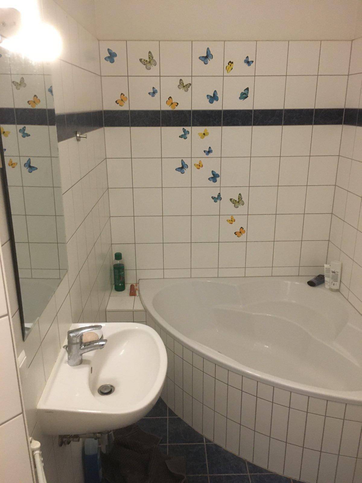 Badezimmer mit Eckbadewanne, Waschbecken und verspielten Schmetterlingsaufklebern an den Fliesen.