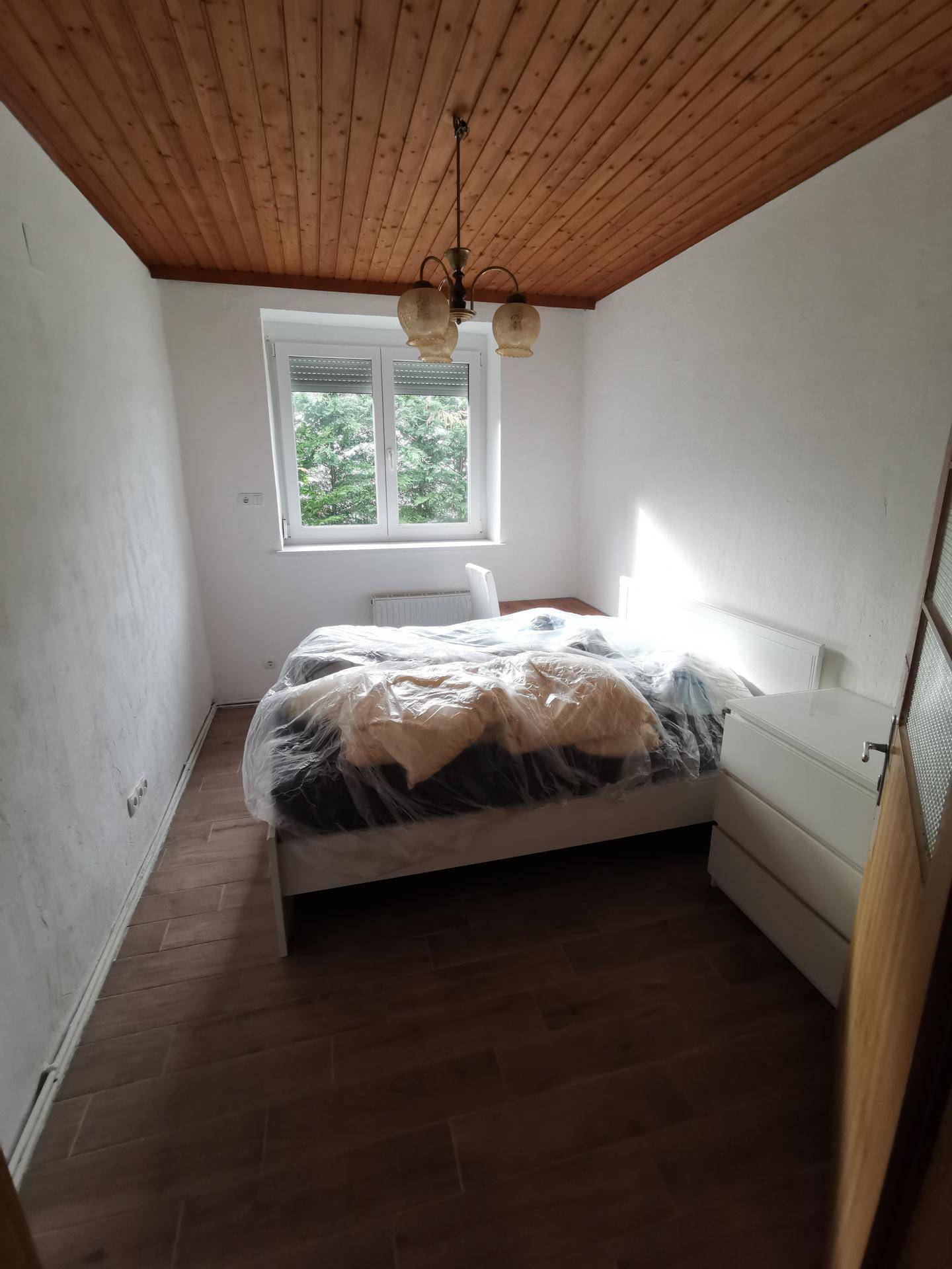 Schlafzimmer mit Holzdecke, Fliesenboden in Holzoptik und einem Fenster mit Blick ins Grüne.