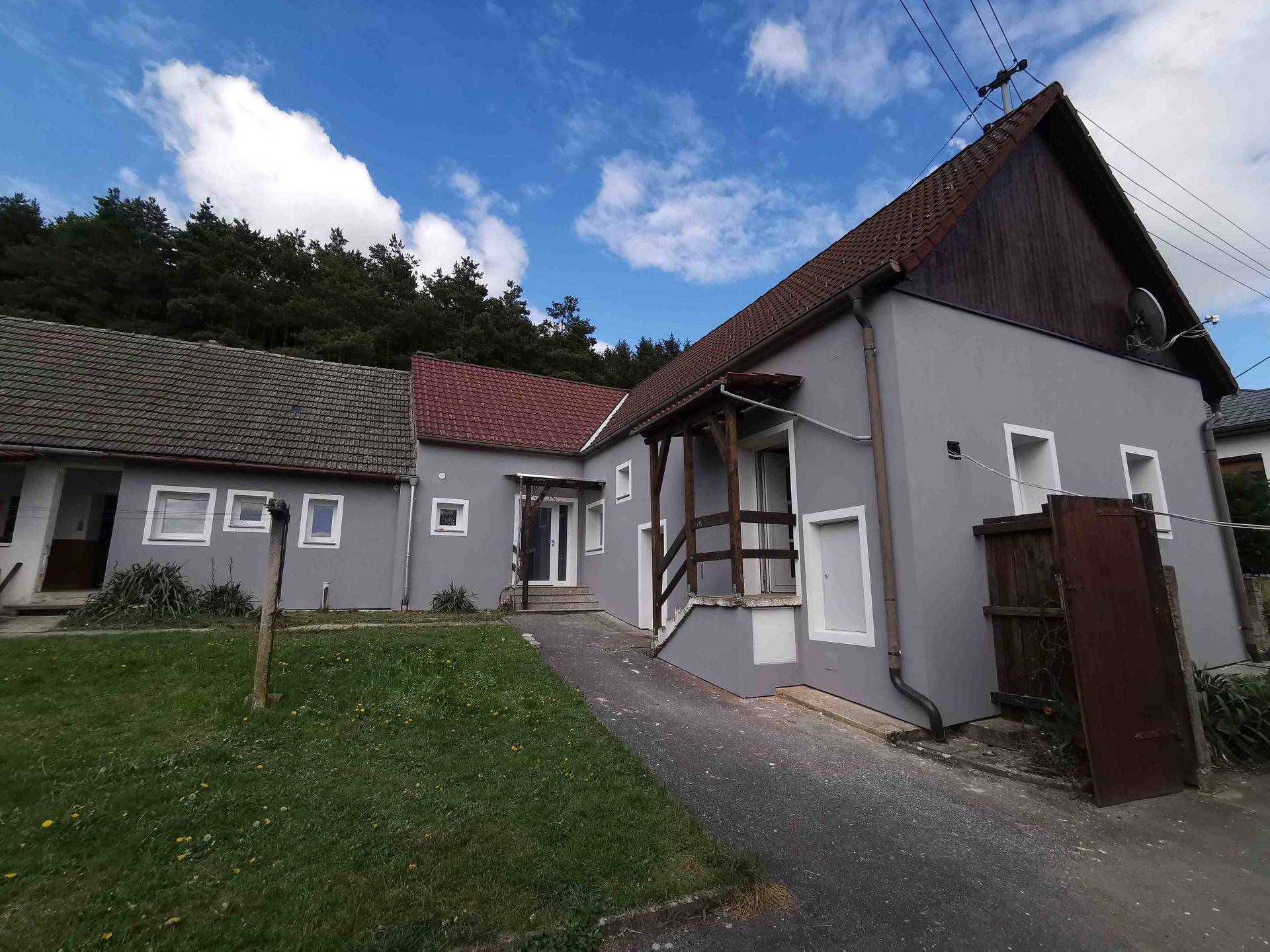 Ansicht des Wohnhauses mit grauer Fassade, Satteldach und angrenzendem Wald im Hintergrund.