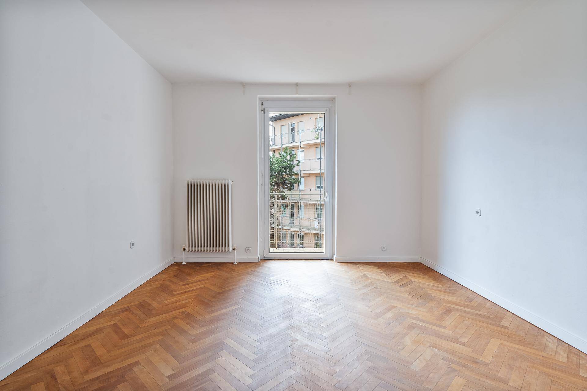 Leeres Zimmer mit Fischgrätparkett und Zugang zu einem Balkon mit Stadtblick.