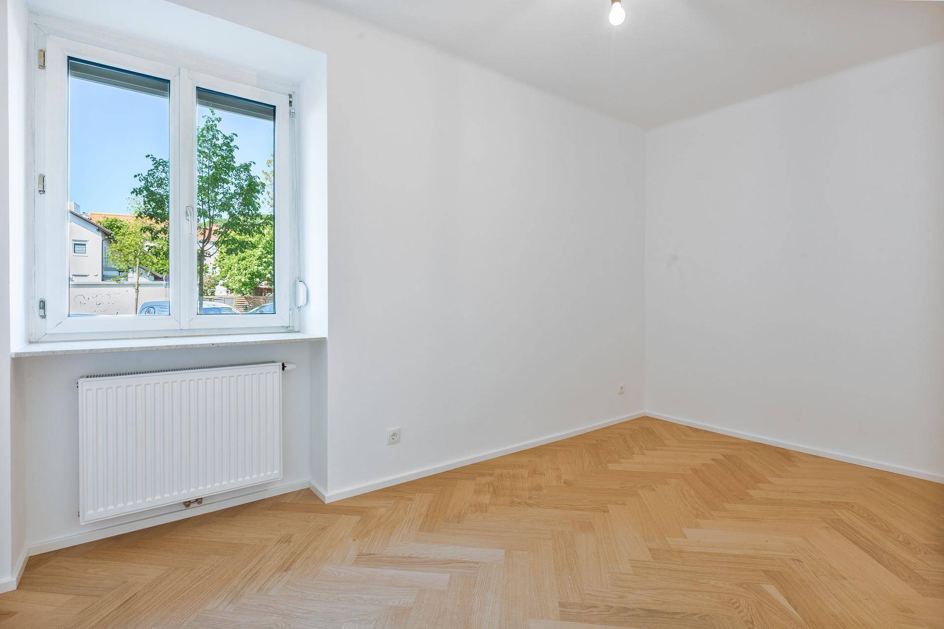 Helles Zimmer mit Fischgrätparkettboden, weißen Wänden und Fenster mit Blick auf die Straße.