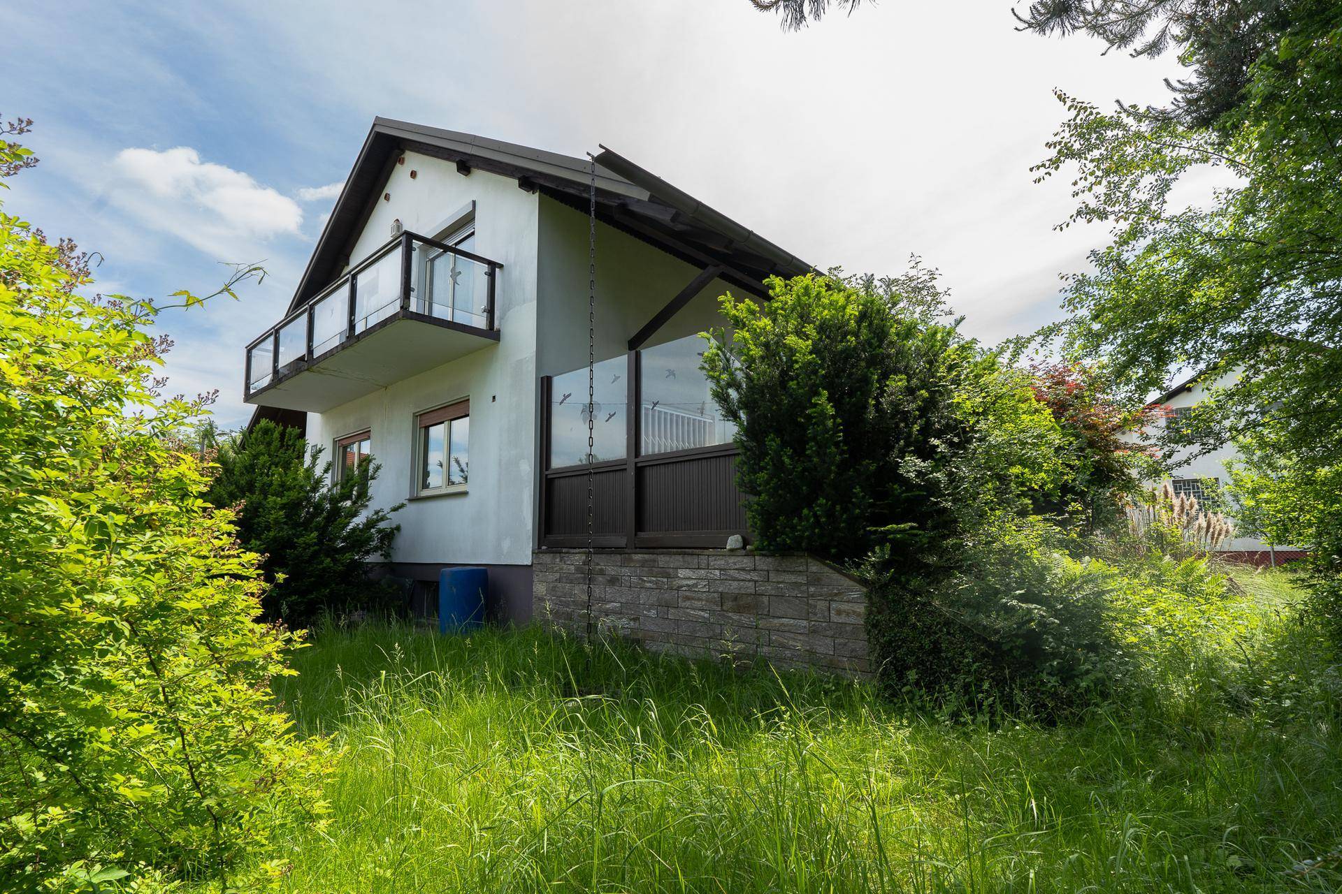 Seitenansicht des Hauses mit Balkon und umgebender grüner Vegetation unter blauem Himmel.