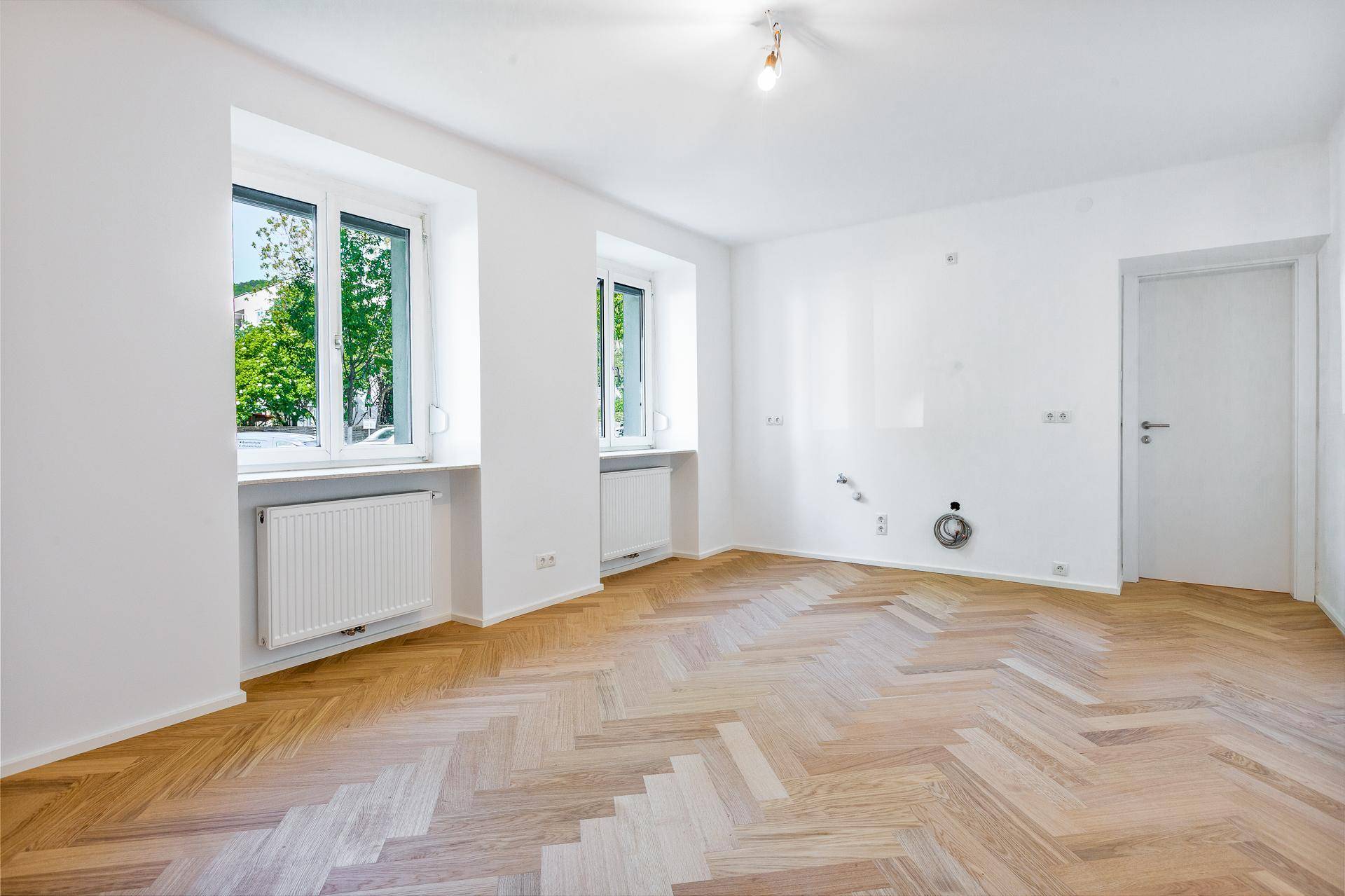 Geräumiges Zimmer mit Fischgrätparkett, weißen Wänden und Anschlüssen für eine mögliche Küche.