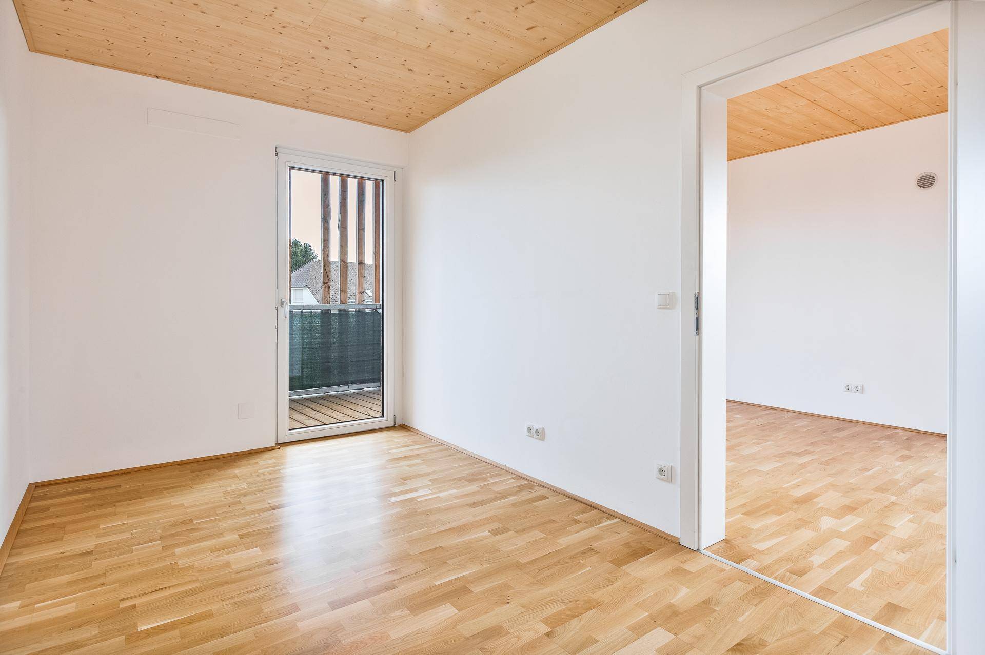 Helles Zimmer mit Holzboden, Holzdecke und Zugang zu einem Balkon.