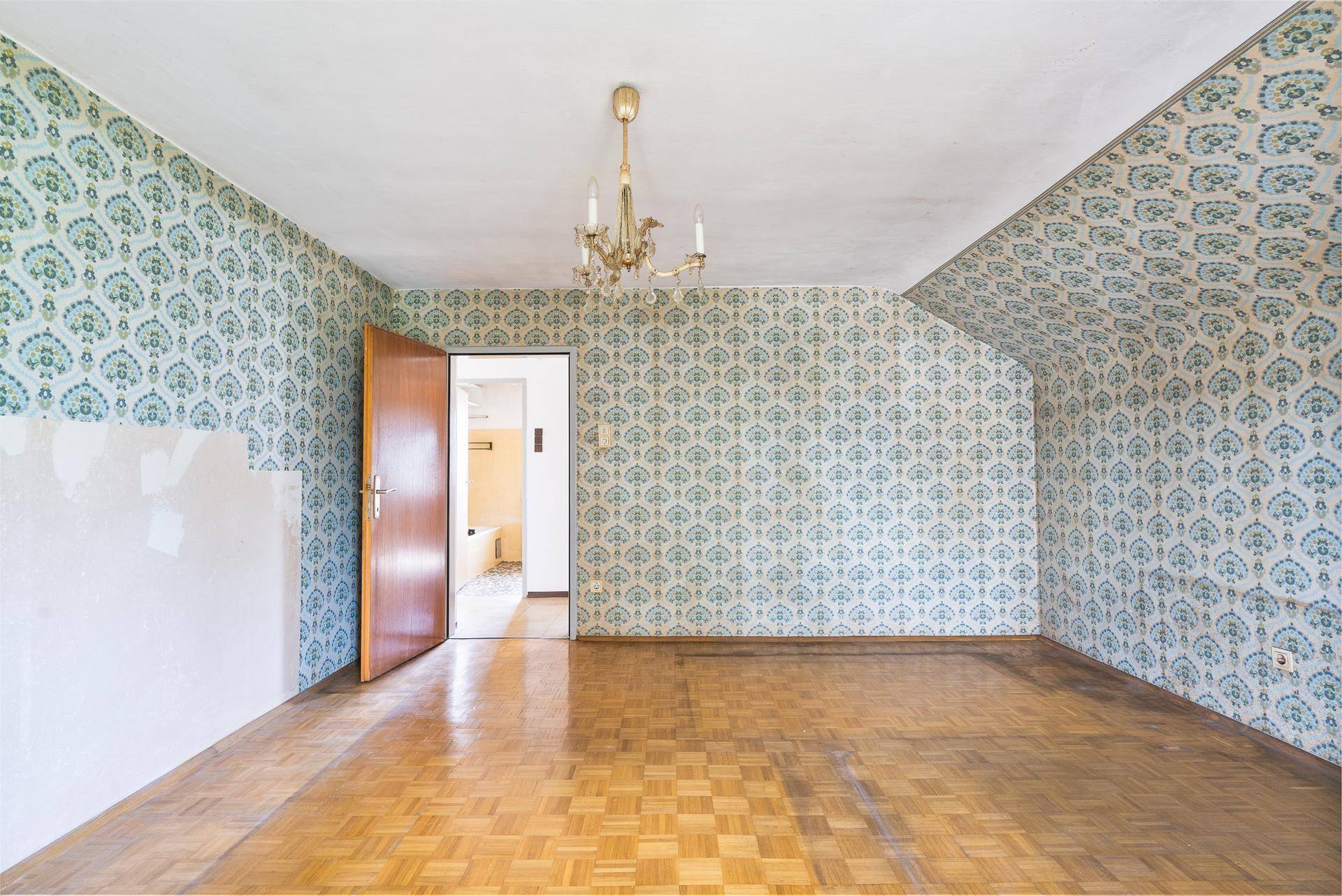 Schlafzimmer mit gemusterter Tapete, Parkettboden und Blick in den Flur.