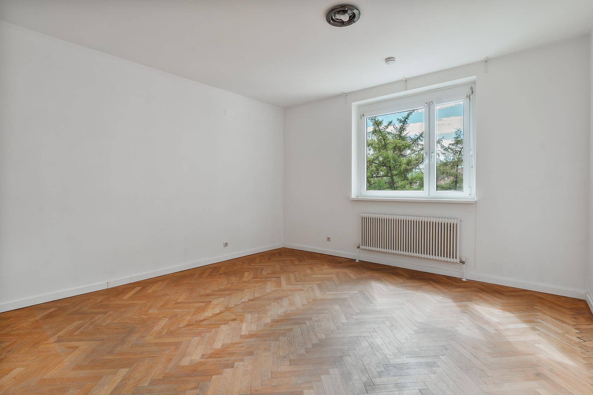 Helles Zimmer mit Fischgrätparkettboden und großem Fenster mit Blick ins Grüne.