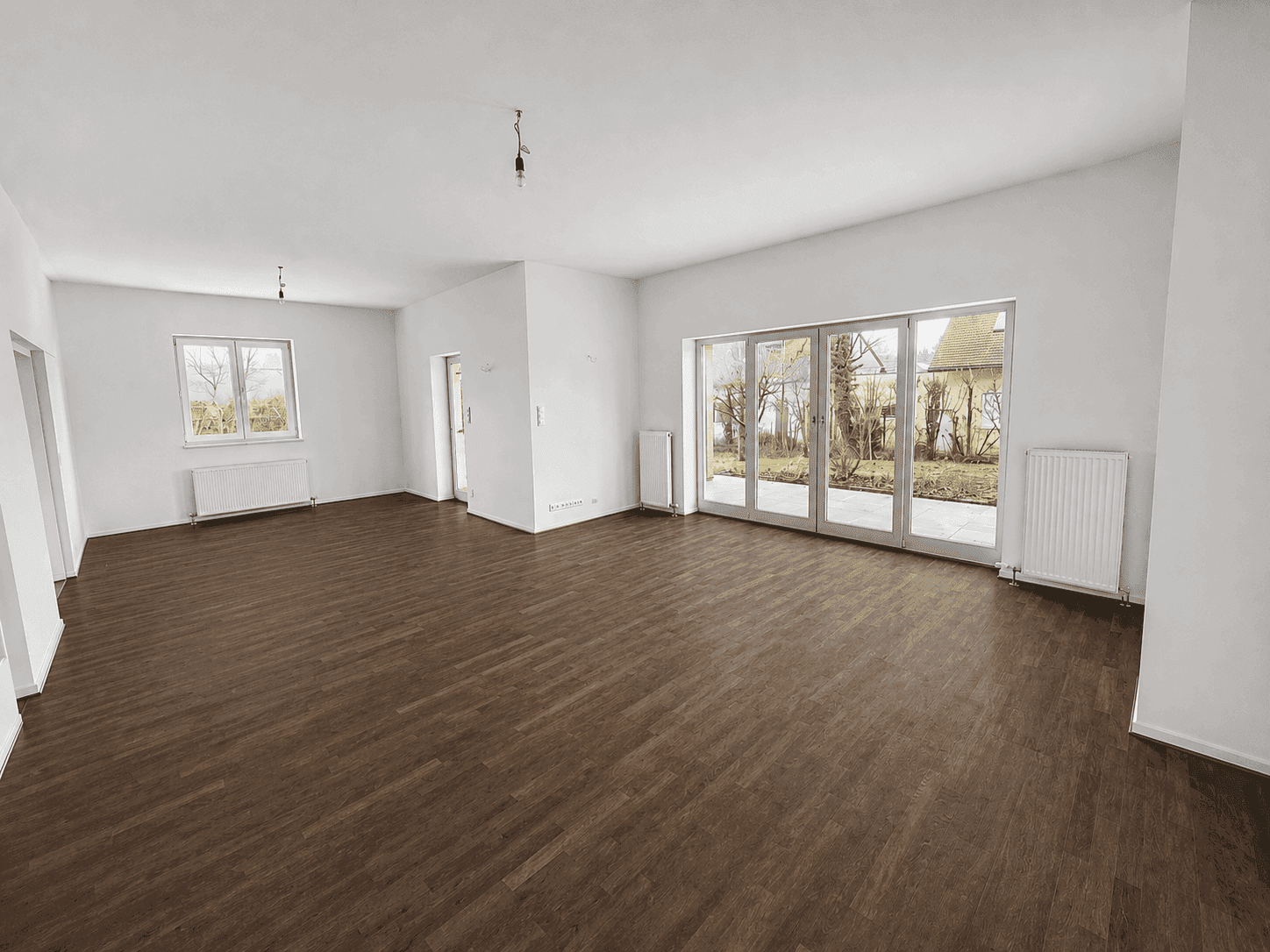 Geräumiges, leeres Wohnzimmer mit dunklem Holzboden und großen Fenstern zum Garten.