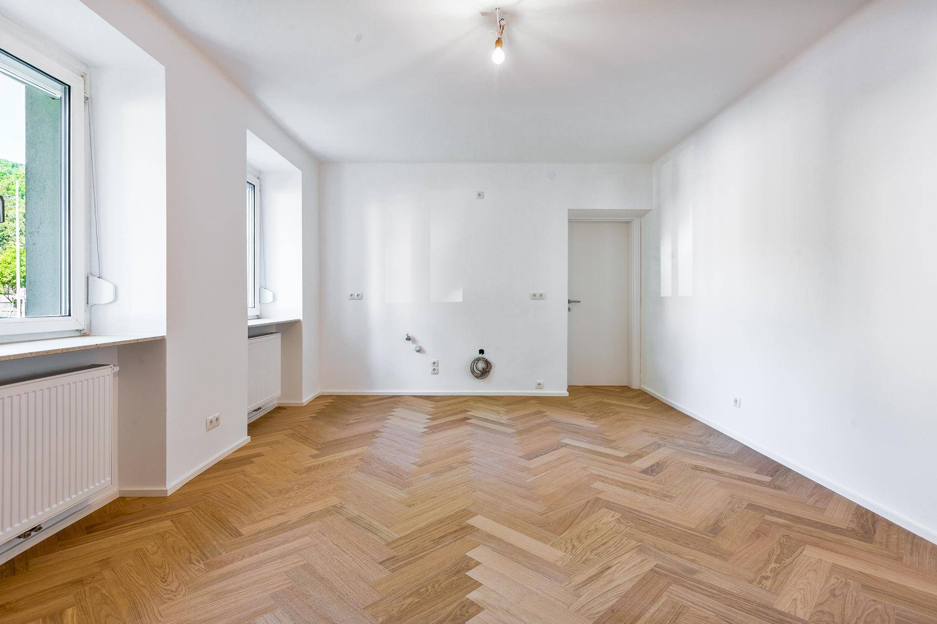 Heller Raum mit Fischgrätparkettboden, weißen Wänden und vorbereiteten Anschlüssen für eine Kücheninstallation.