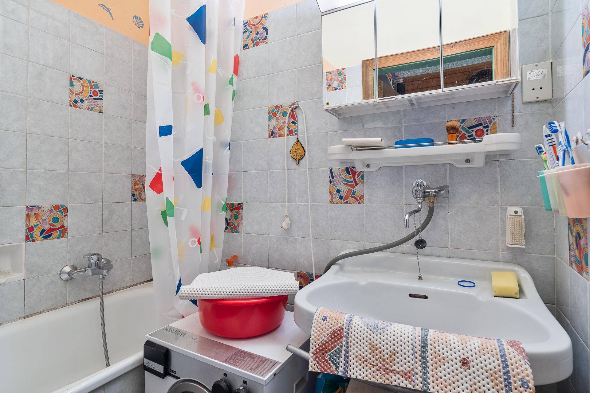Badezimmer mit Badewanne, Duschvorhang und bunten Fliesen im 80er-Jahre-Stil.