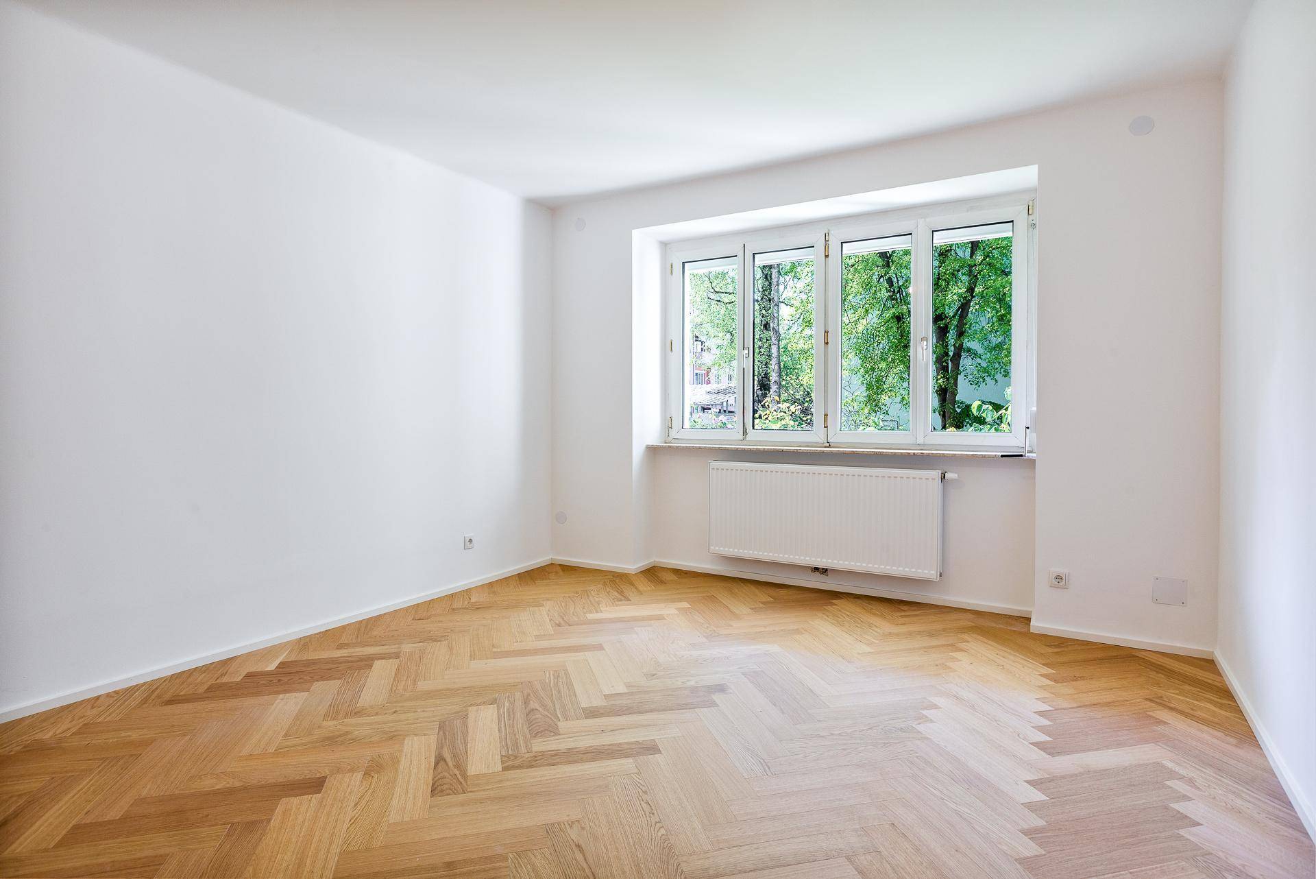 Lichtdurchflutetes Zimmer mit Fischgrätparkett, weißen Wänden und einem großen Fenster mit Grünblick.