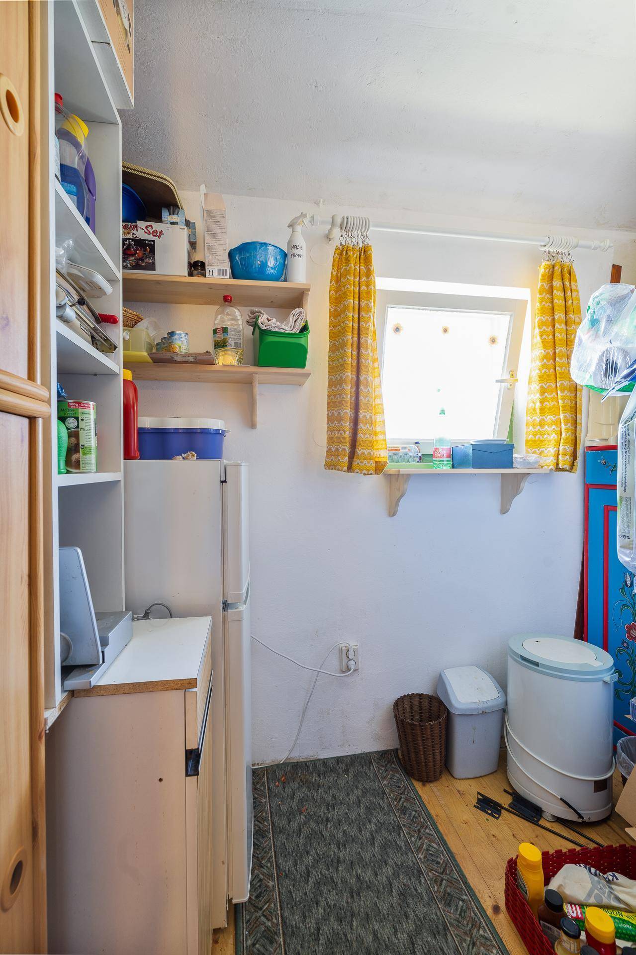 Praktischer Abstellraum mit Regalen, Kühlschrank und einem Fenster mit gelben Vorhängen.