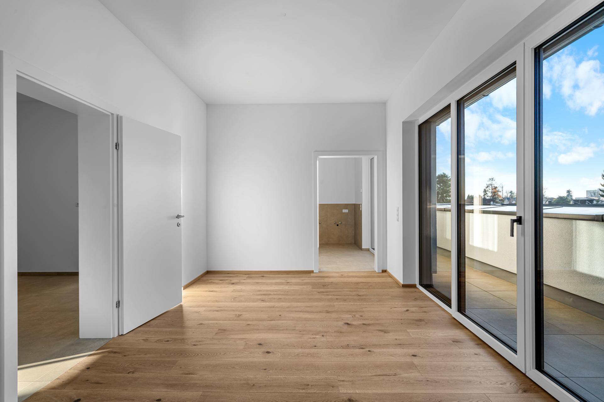 Geräumiger Raum mit hellem Holzparkett und Zugang zu weiteren Zimmern und einem Balkon.
