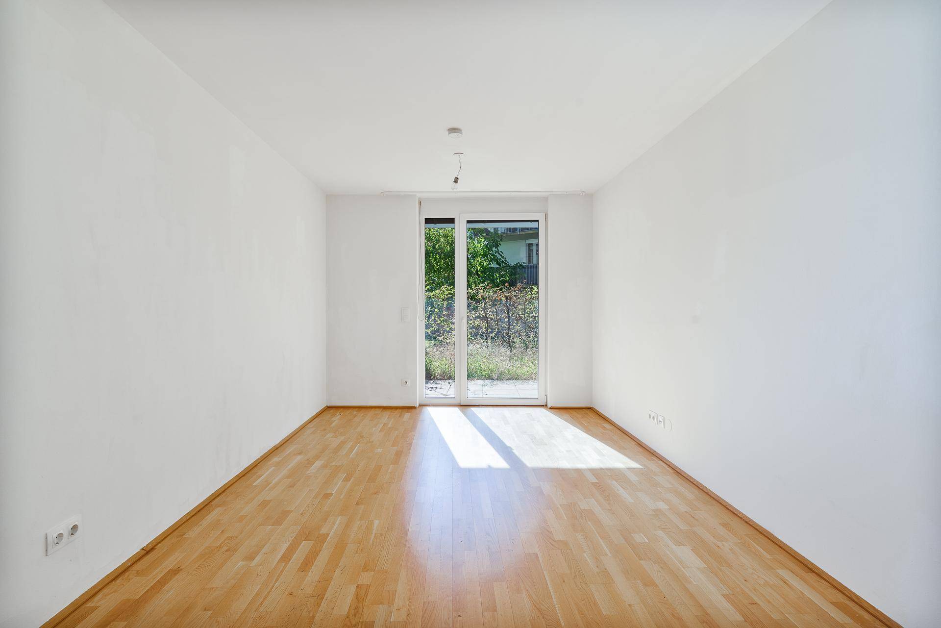 Leerer, heller Raum mit Holzboden und großer Glastür, die zu einem Außenbereich führt.