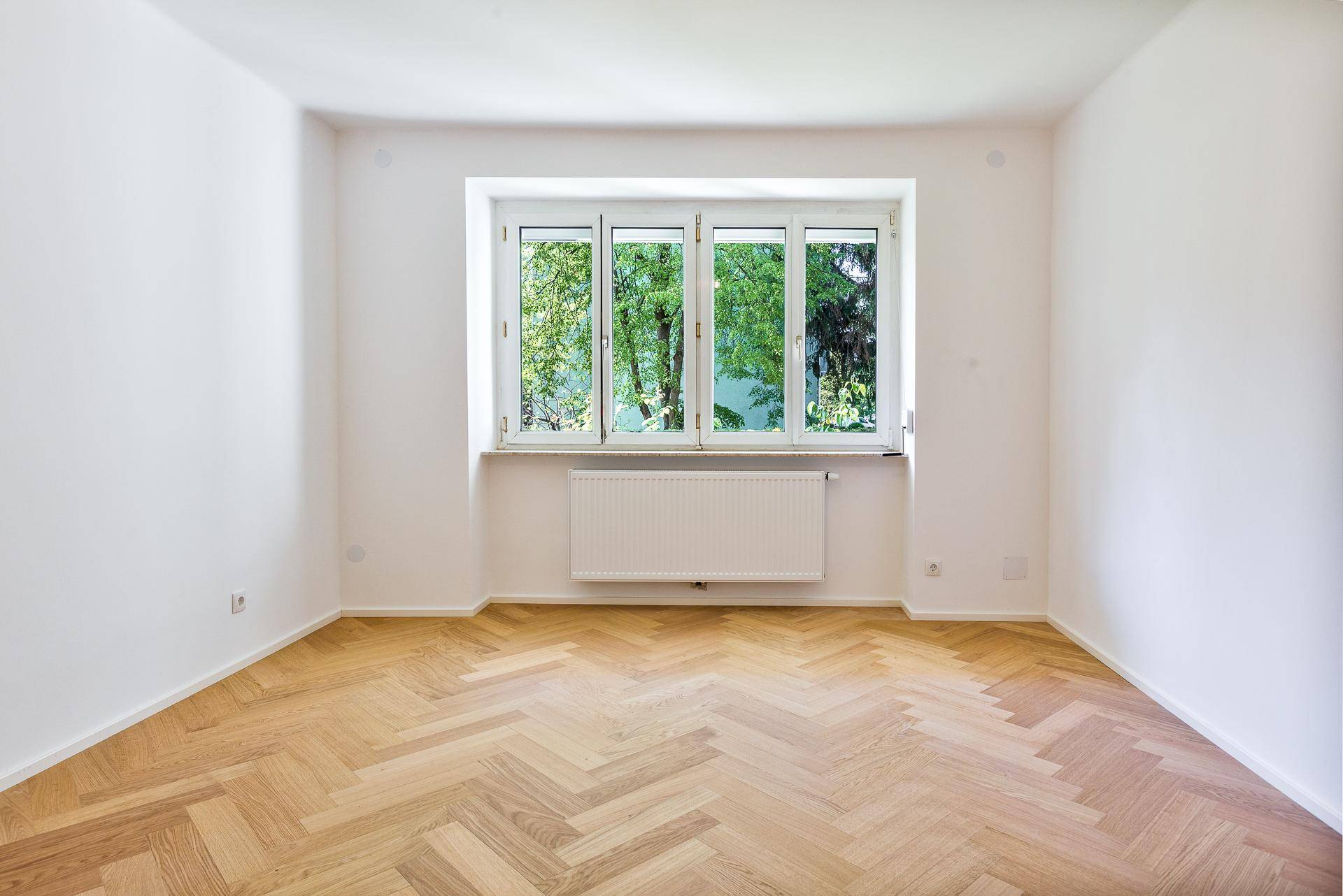 Geräumiges Zimmer mit Fischgrätparkettboden, weißen Wänden und einem breiten Fenster mit Blick ins Grüne.
