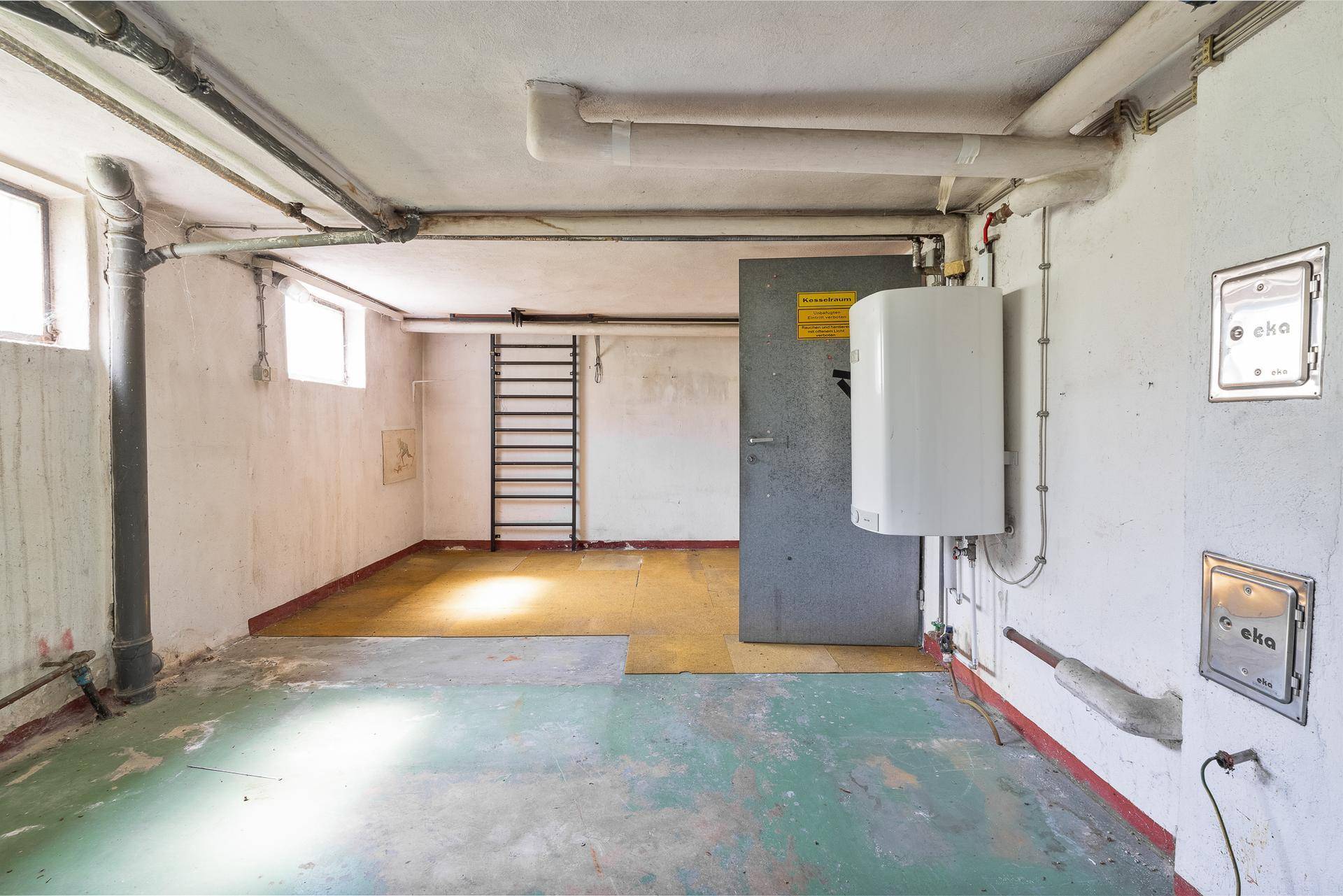 Kellerraum mit Warmwasserbereiter, sichtbaren Leitungen und Fenstern.