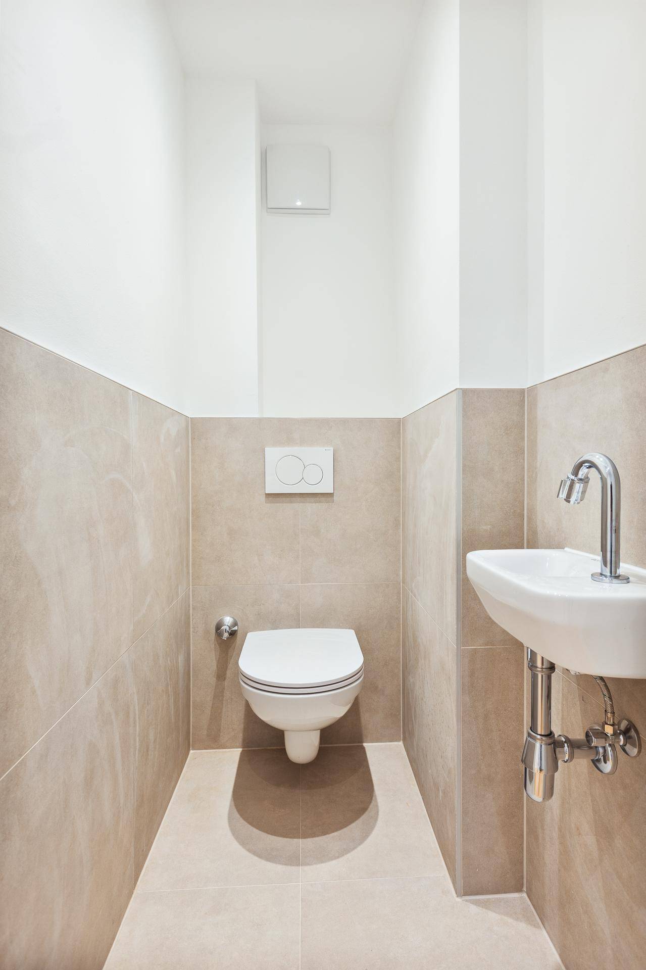 Modernes Gäste-WC mit wandhängender Toilette, kleinem Waschbecken und beigefarbenen Fliesen.