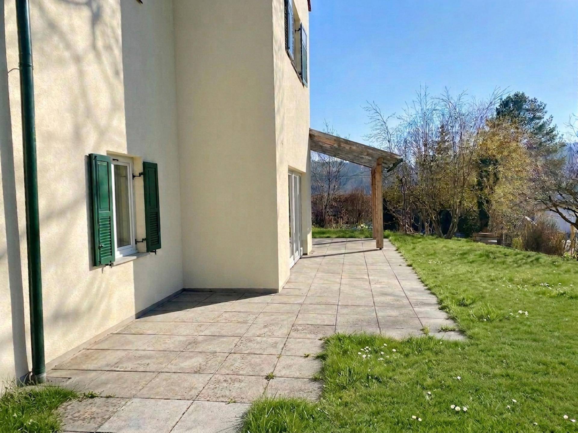 Geräumige gepflasterte Terrasse neben dem Haus, umgeben von Rasen und Bäumen.