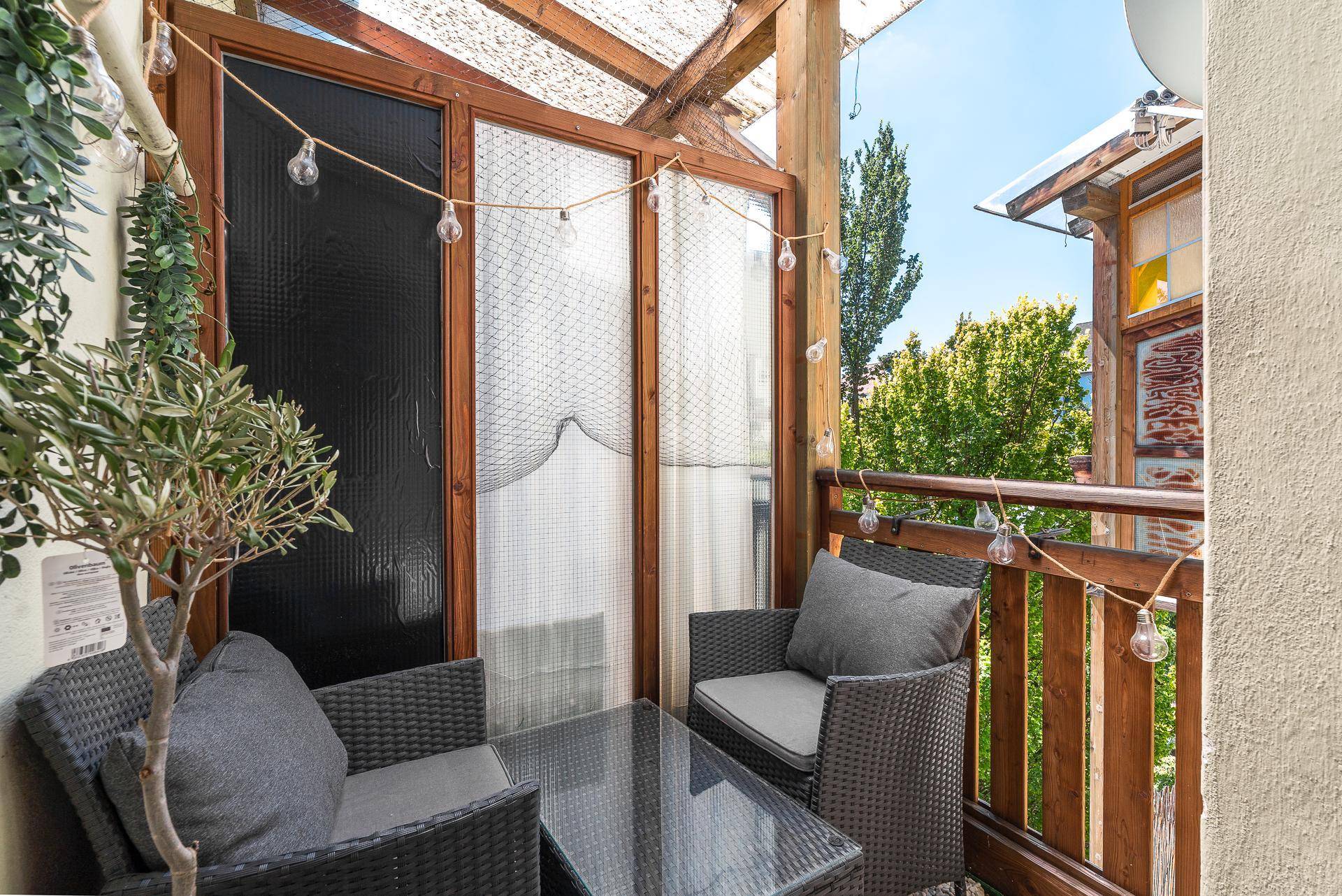 Überdachter Balkon mit Rattanmöbeln und Lichterkette, umgeben von viel Grün.