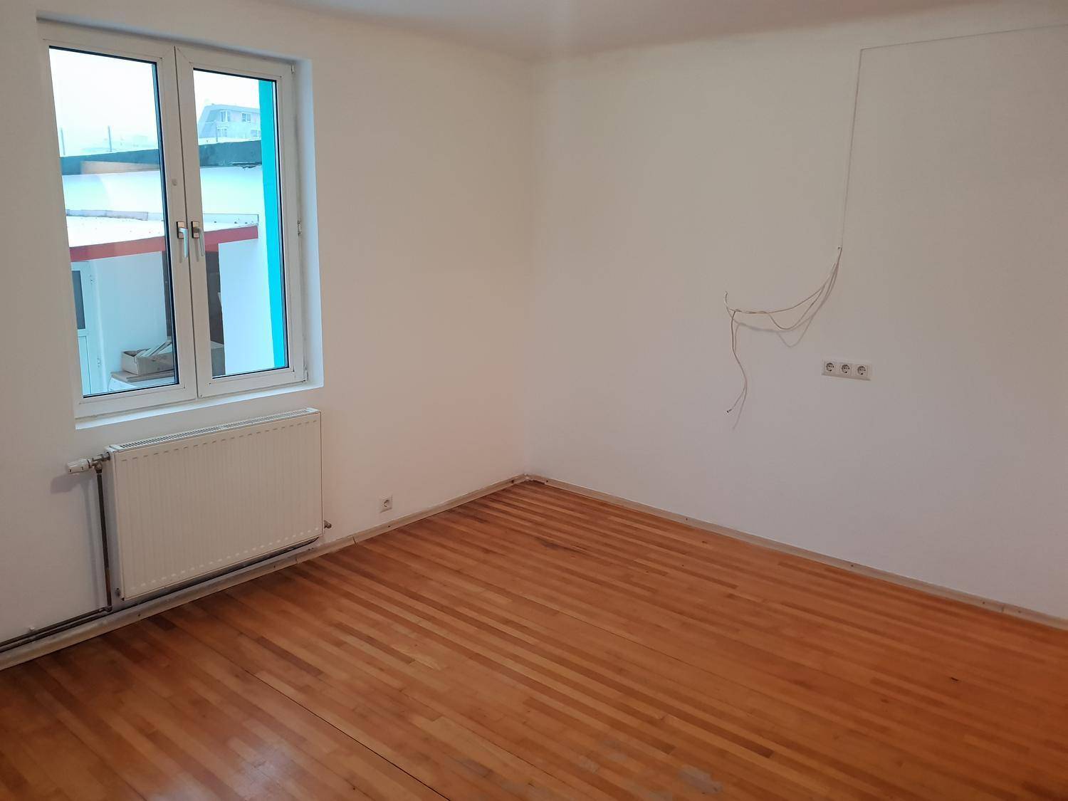 Leerer Raum mit Holzboden und Fenster, unfertige Verkabelung an der Wand sichtbar.
