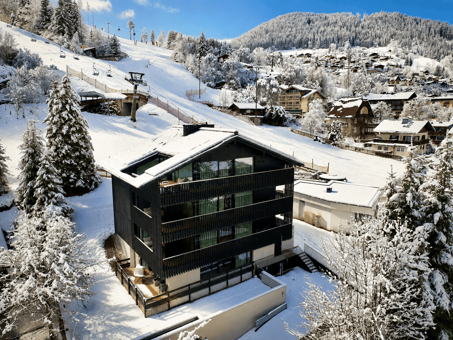 Luftaufnahme des modernen Gebäudes in verschneiter Winterlandschaft, mit Skipiste im Hintergrund.
