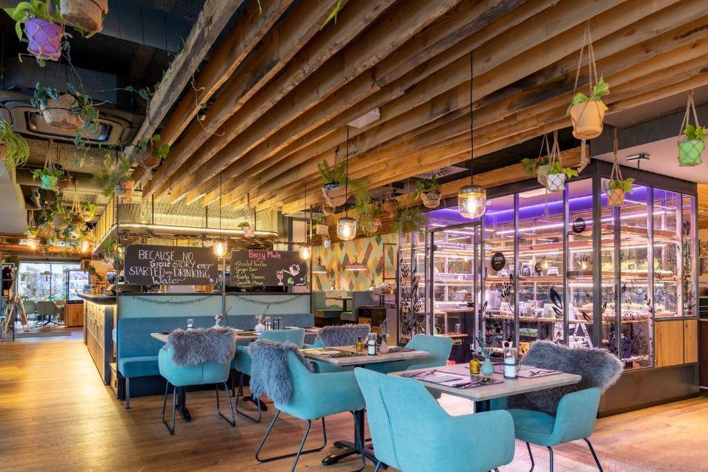 Modernes Restaurant mit Holzbalkendecke, farbigen Stühlen und hängenden Pflanzen.