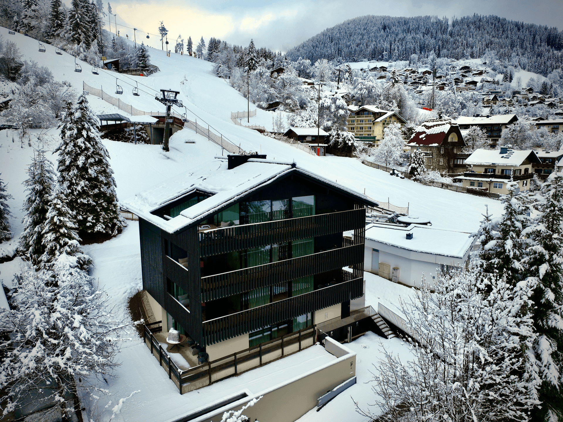 Luftaufnahme des modernen Gebäudes in verschneiter Berglandschaft, nahe einer Skipiste gelegen.