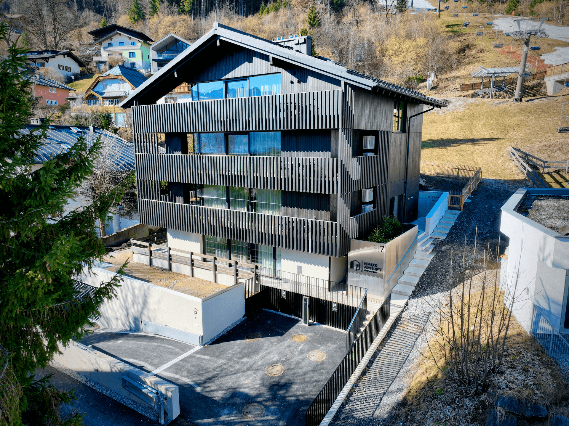 Außenansicht des modernen Gebäudes mit dunkler Fassade und Balkonen, im Hintergrund eine Skipiste.