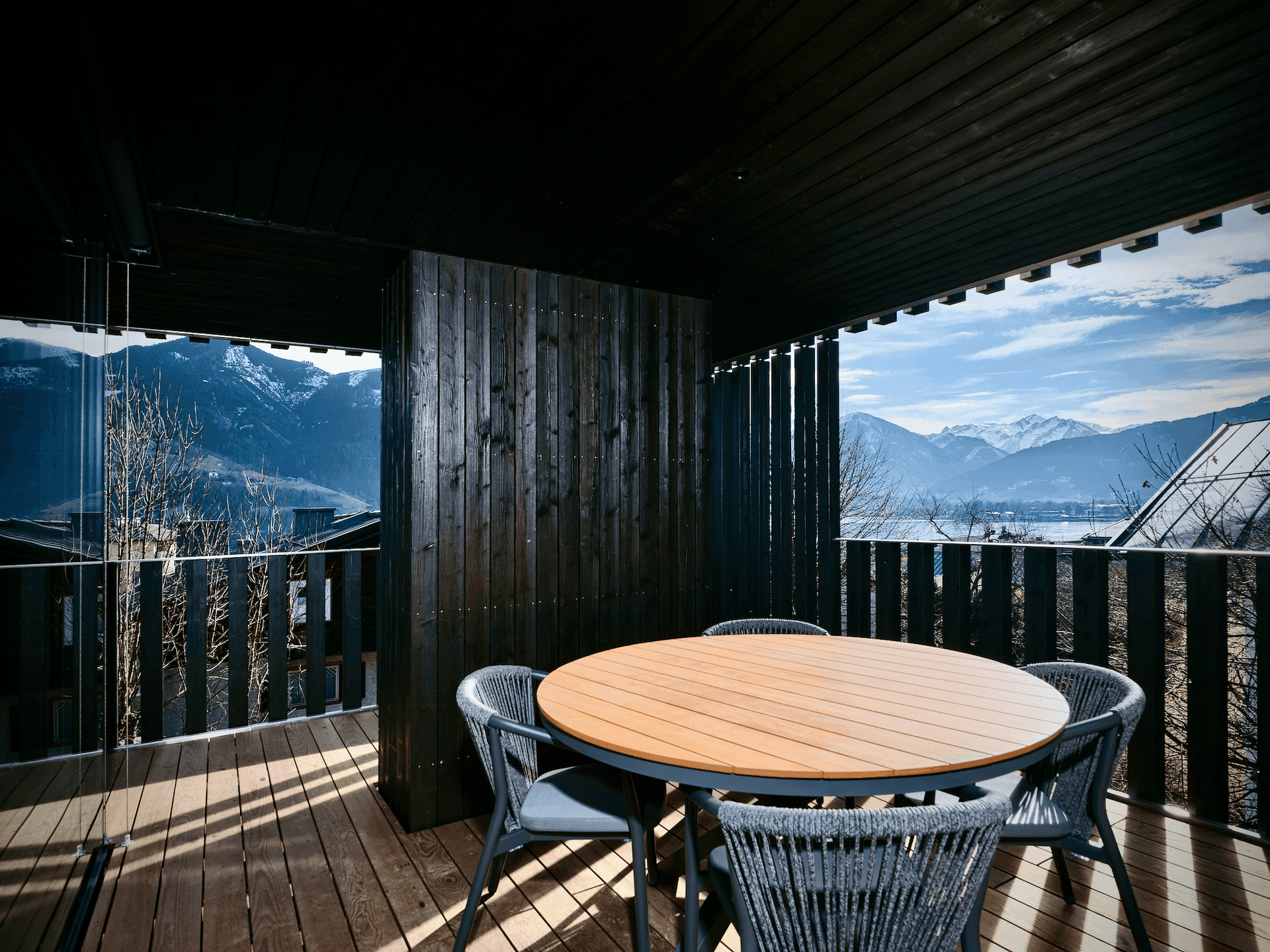 Gemütlicher Balkon mit Holztisch und Stühlen, umgeben von dunklen Holzlamellen, mit Bergblick.