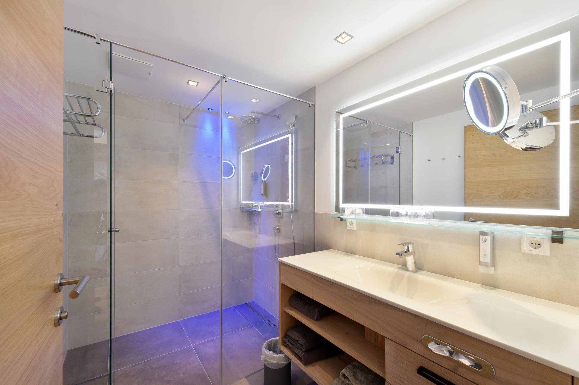 Modernes Badezimmer mit begehbarer Dusche, beleuchtetem Spiegel und Holzwaschtisch.