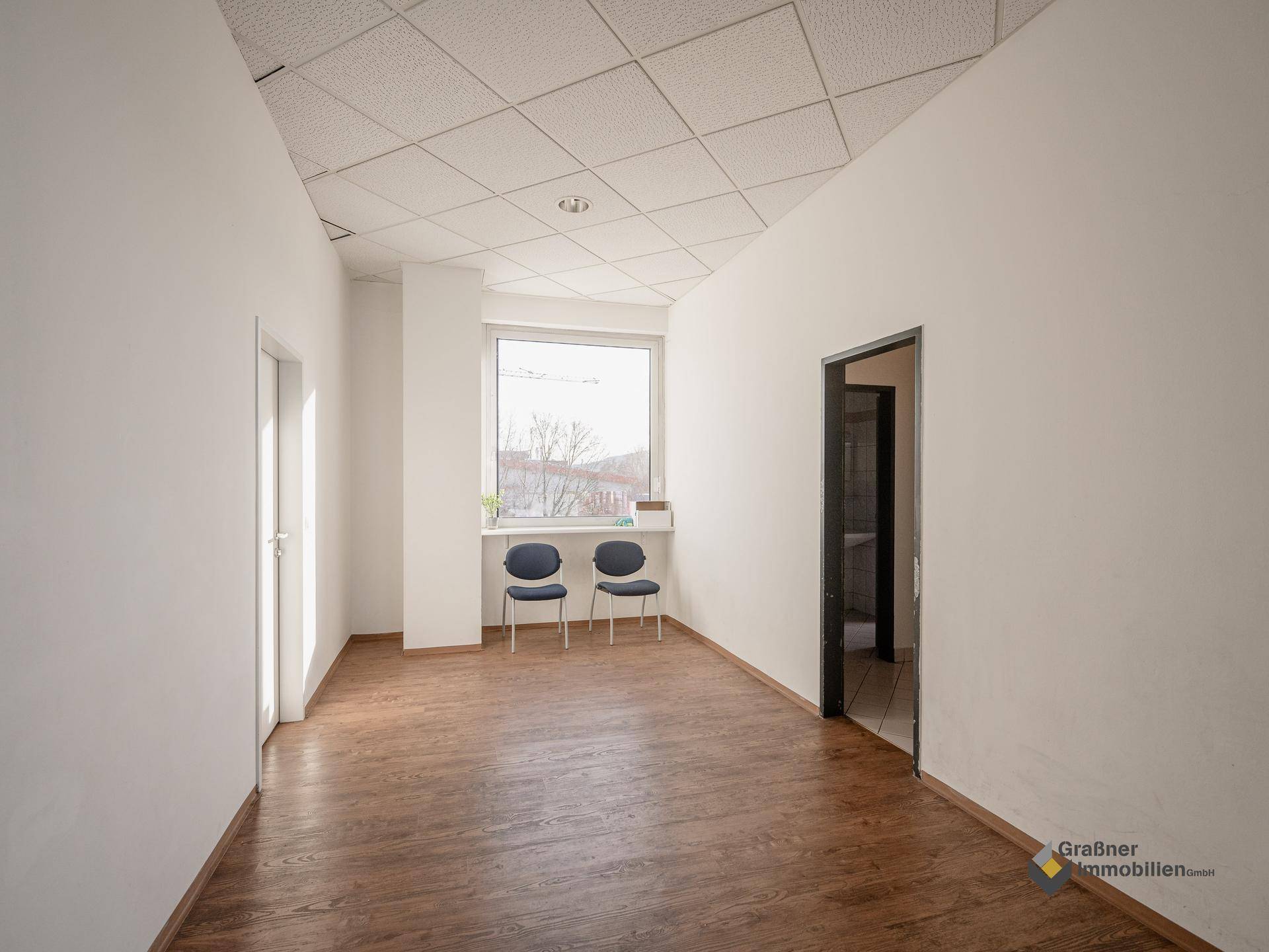 Leerer Büroraum mit Laminatboden, weißen Wänden und einem Fenster, das Tageslicht hereinlässt.