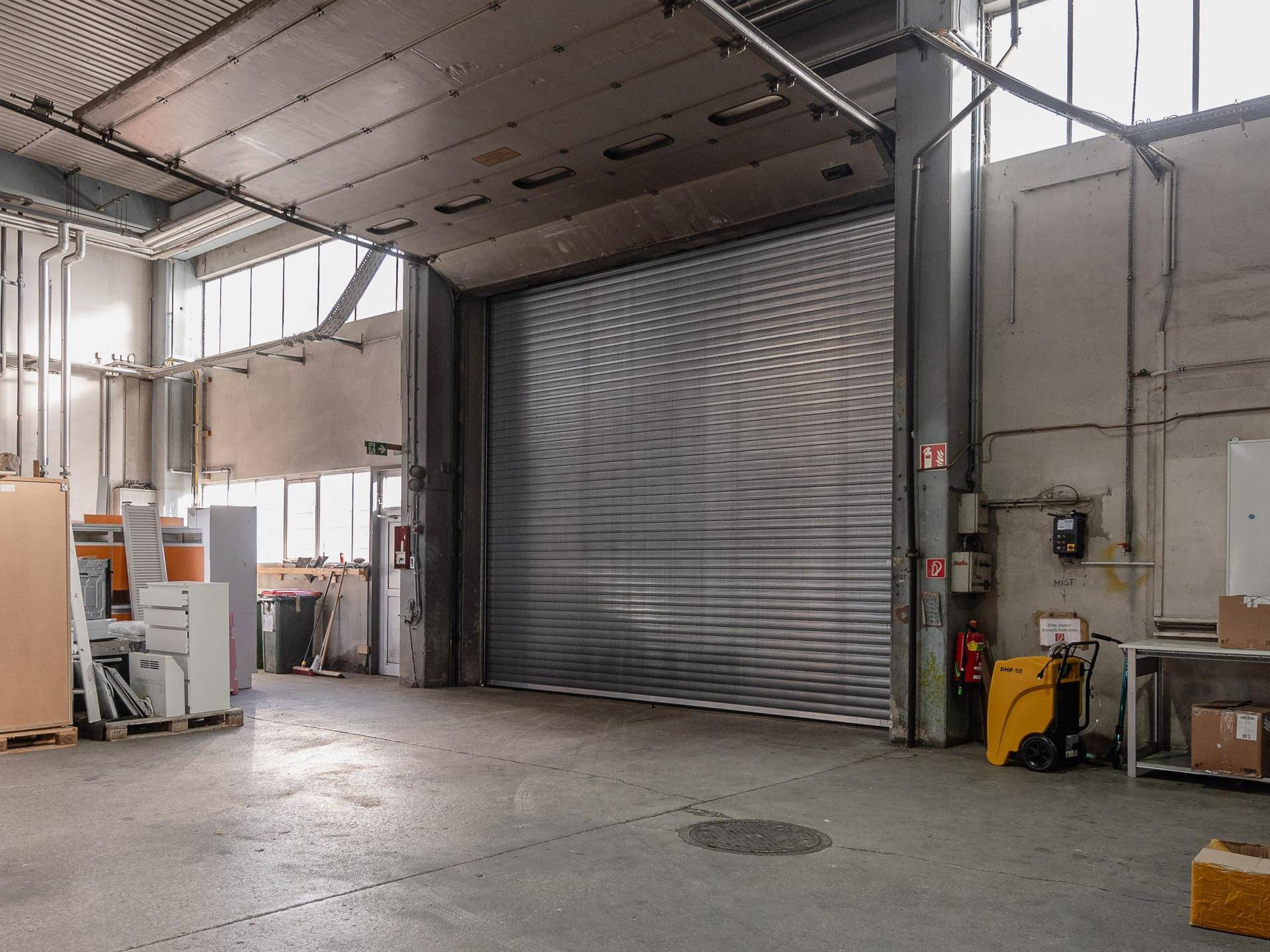 Geräumige Lagerhalle mit großem Rolltor, Betonboden und sichtbaren Installationen an der Decke.