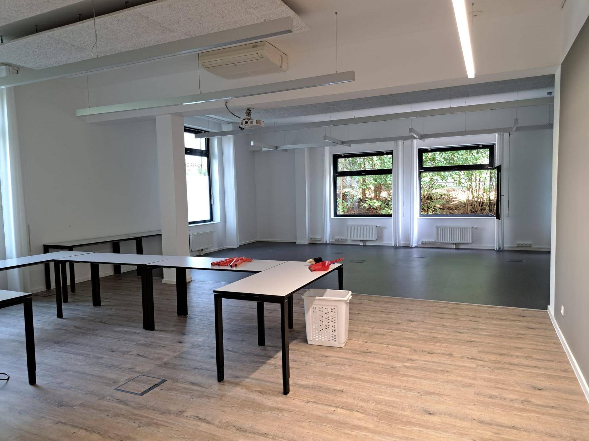 Geräumiger Mehrzweckraum mit flexiblen Tischen, großen Fenstern und Blick ins Grüne. Die Decke ist mit Akustikplatten ausgestattet.