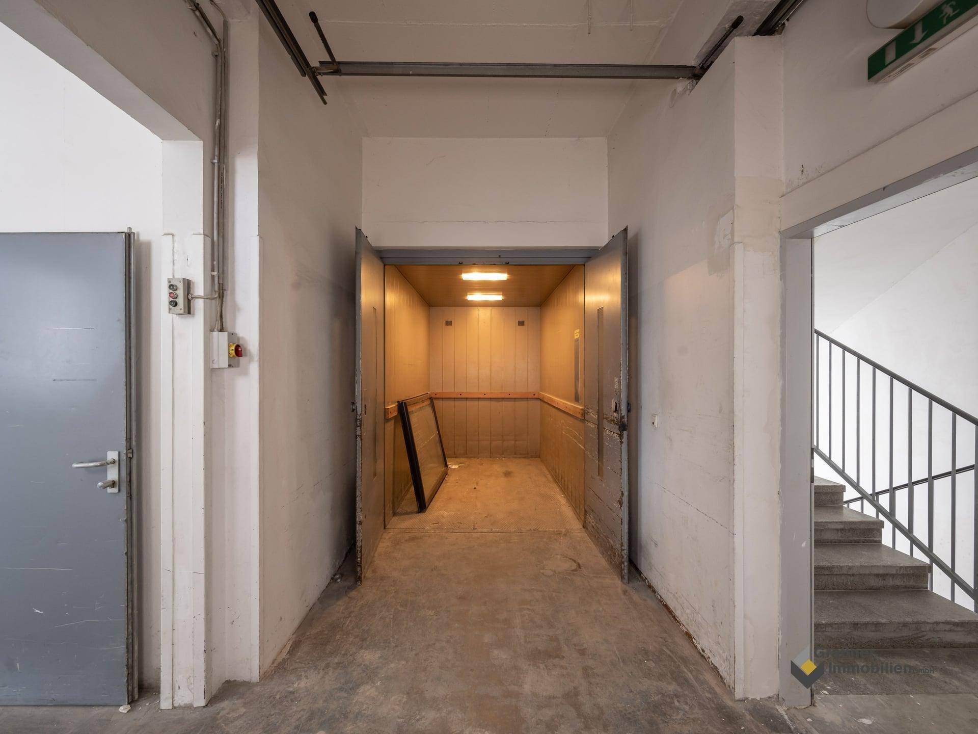 Flur mit offenem Lastenaufzug und angrenzendem Treppenhaus, alles in funktionalem Zustand.